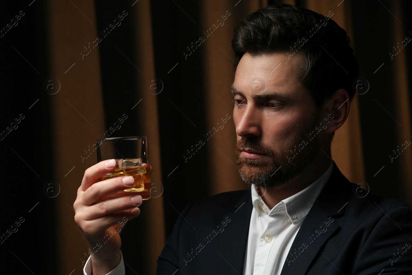 Photo of Man in suit holding glass of whiskey with ice cubes on brown background