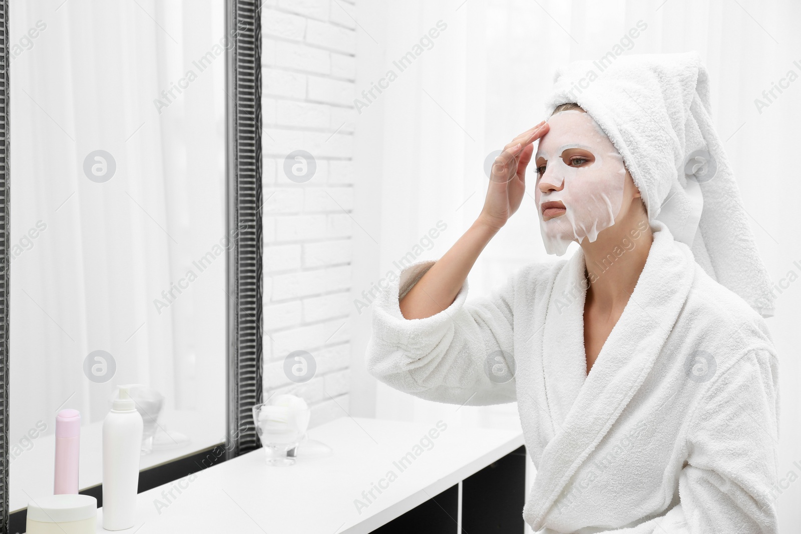 Photo of Beautiful woman with sheet mask on her face at mirror indoors