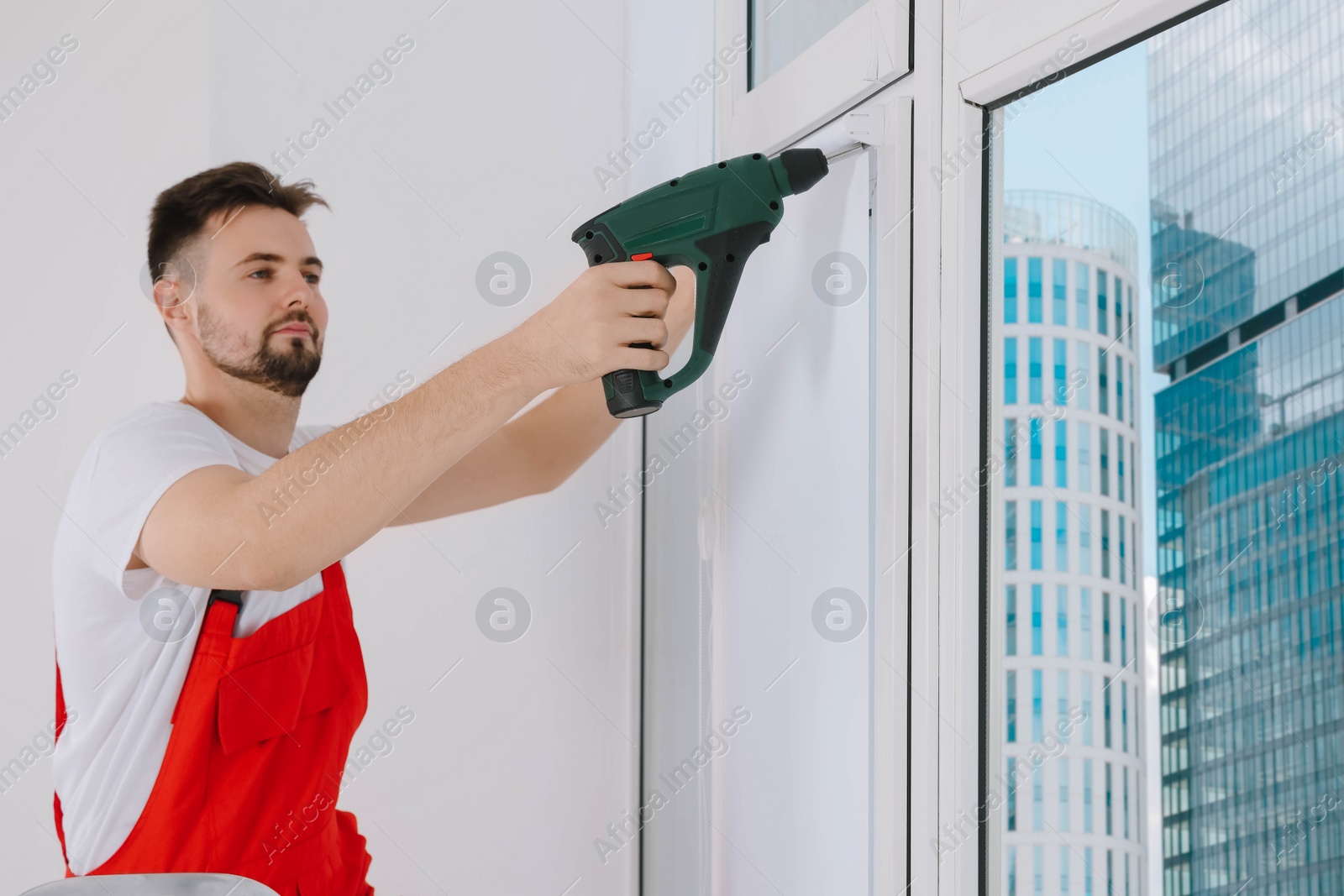Photo of Worker in uniform installing roller window blind indoors, focus on drill