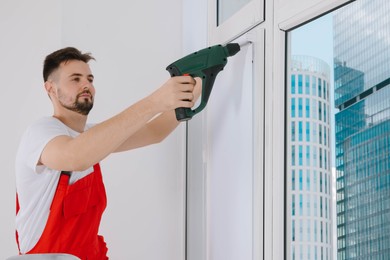 Photo of Worker in uniform installing roller window blind indoors, focus on drill