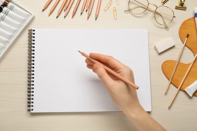 Photo of Woman with pencil and blank sketchbook at white wooden table, top view