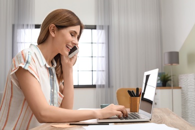 Photo of Young woman talking on phone while working with laptop at desk in home office