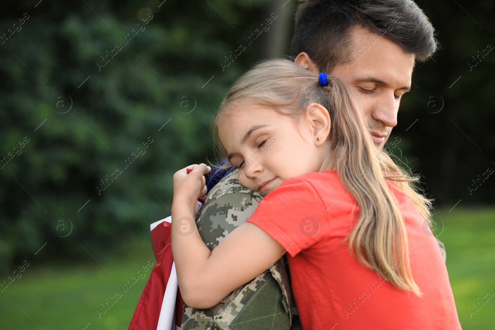 Photo of Father in military uniform with American flag hugging his little daughter at green park