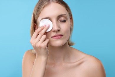 Young woman cleaning face with cotton pad on light blue background