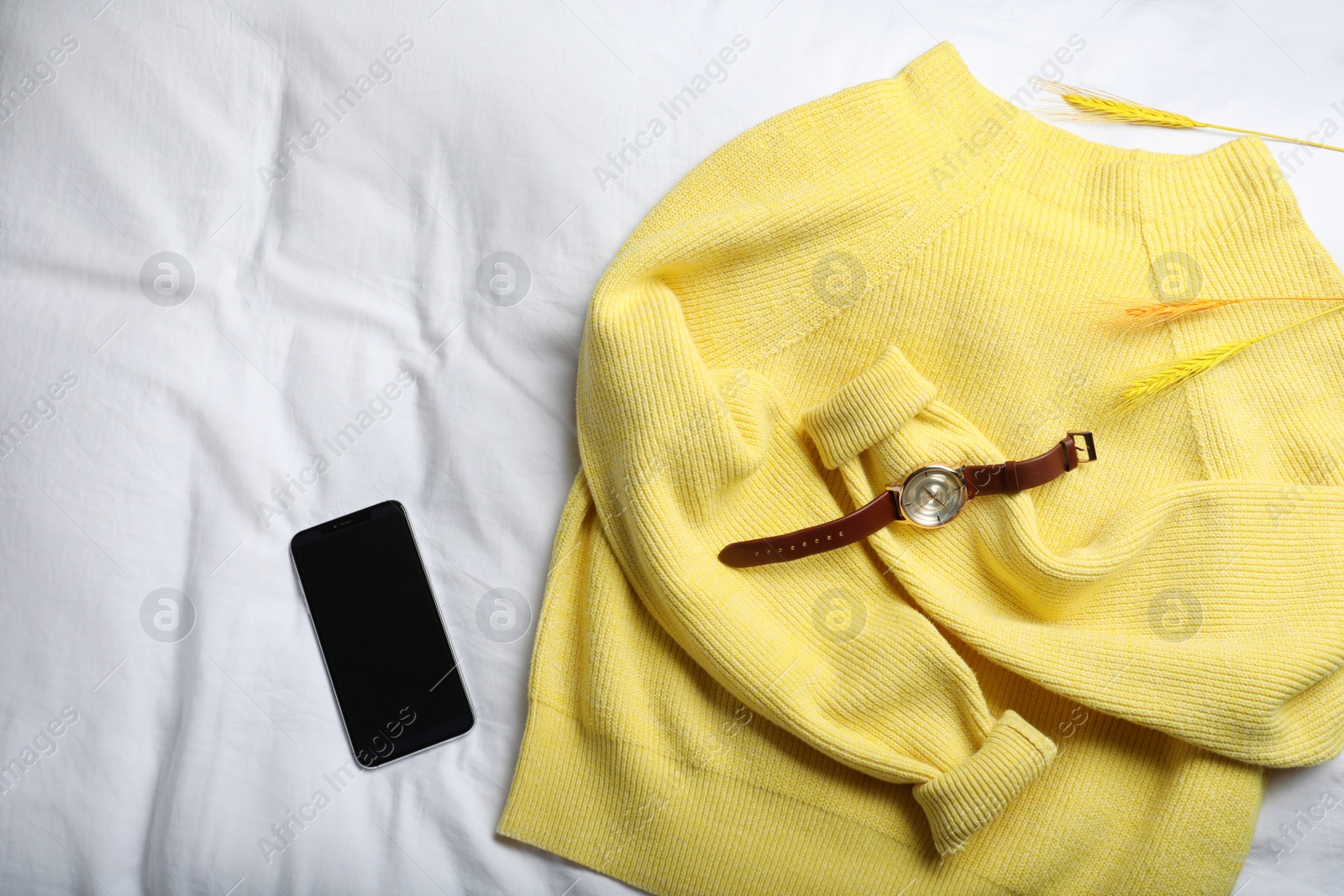 Photo of Yellow warm sweater with smartphone and wristwatch on bedsheet, flat lay