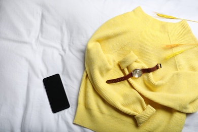 Photo of Yellow warm sweater with smartphone and wristwatch on bedsheet, flat lay