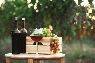 Photo of Bottles and glass of red wine with fresh grapes on wooden table in vineyard