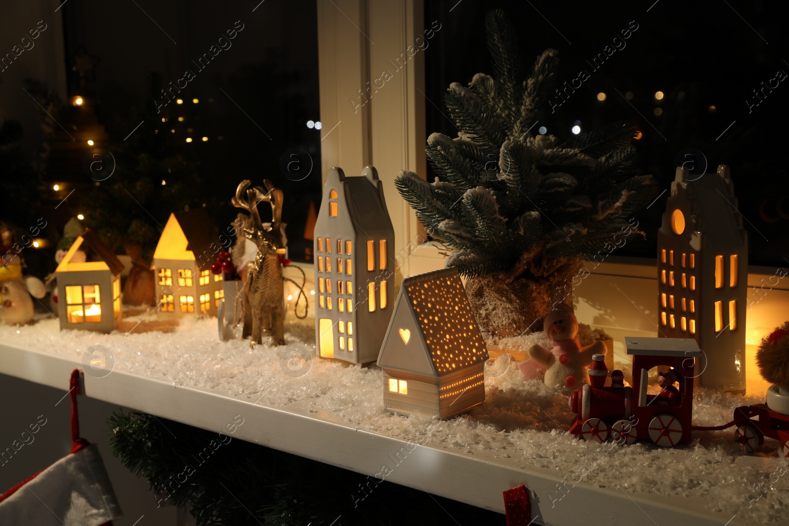 Photo of Christmas atmosphere. Beautiful glowing houses, fir trees, artificial snow and toys on window sill indoors