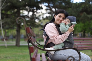 Photo of Mother holding her child in sling (baby carrier) on bench in park