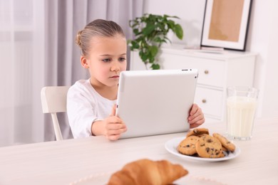 Photo of Little girl using tablet while having breakfast at table indoors. Internet addiction