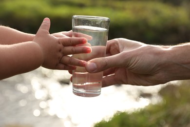 Photo of Father giving her daughter glass of fresh water near stream on sunny day, closeup