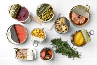 Open tin cans with different products and rosemary on white tiled table, flat lay
