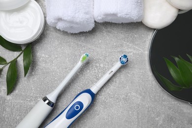 Photo of Electric toothbrushes, rolled towels and green leaves on grey textured table, flat lay