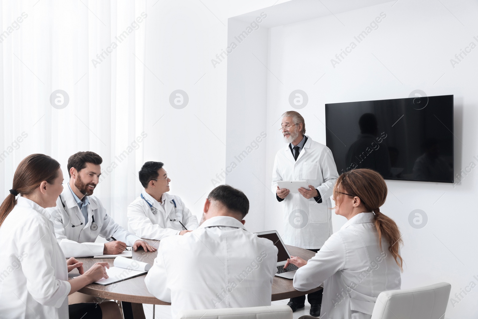 Photo of Team of doctors listening to senior speaker report near tv screen in meeting room. Medical conference