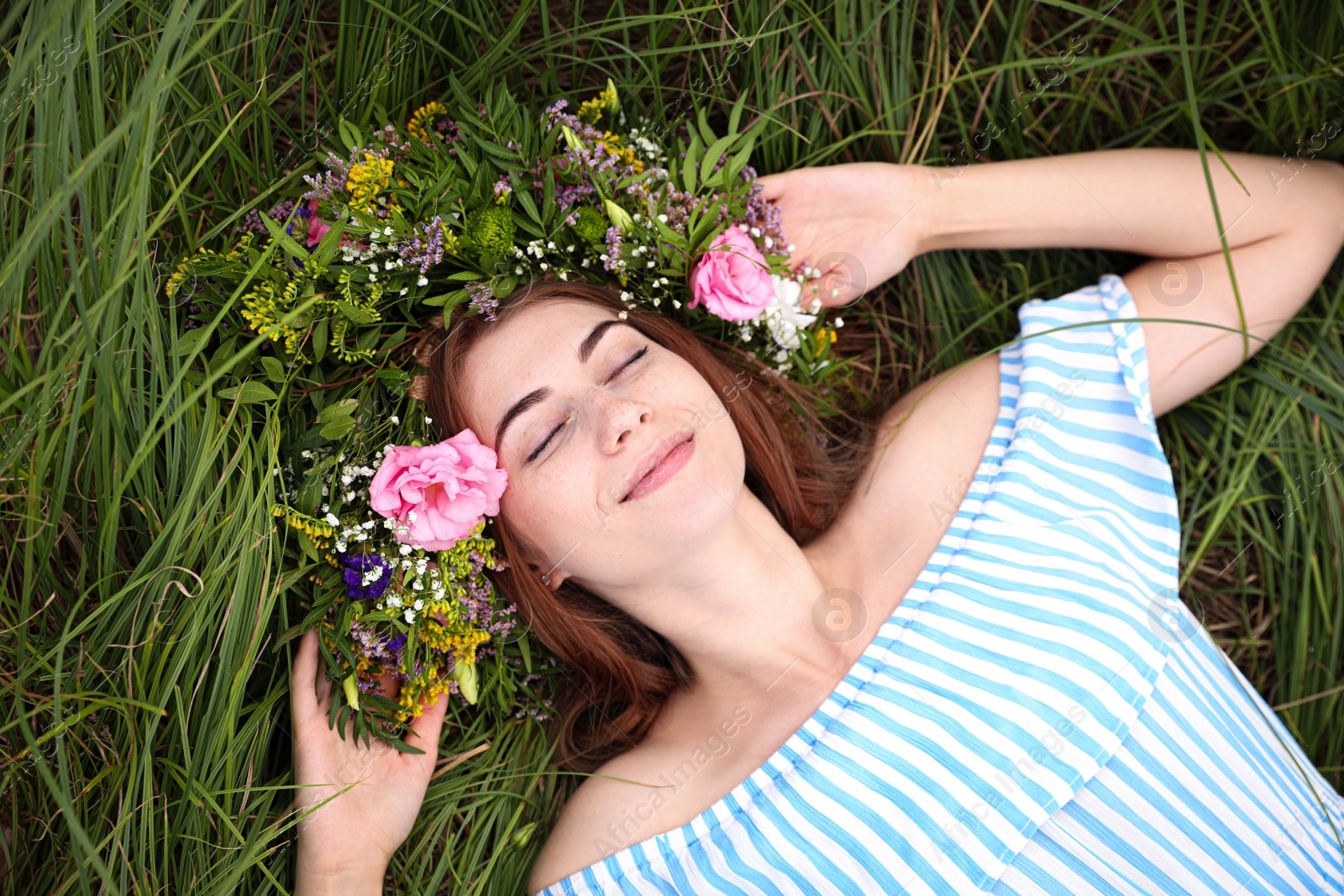 Photo of Young woman wearing wreath made of beautiful flowers on green grass, top view