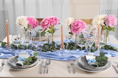 Photo of Beautiful table setting. Plates with greeting cards, napkins and branches near glasses, peonies, burning candles and cutlery on table indoors