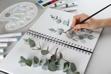 Photo of Woman drawing beautiful eucalyptus branches in sketchbook at white table, closeup