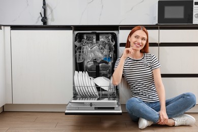 Smiling woman sitting near open dishwasher in kitchen