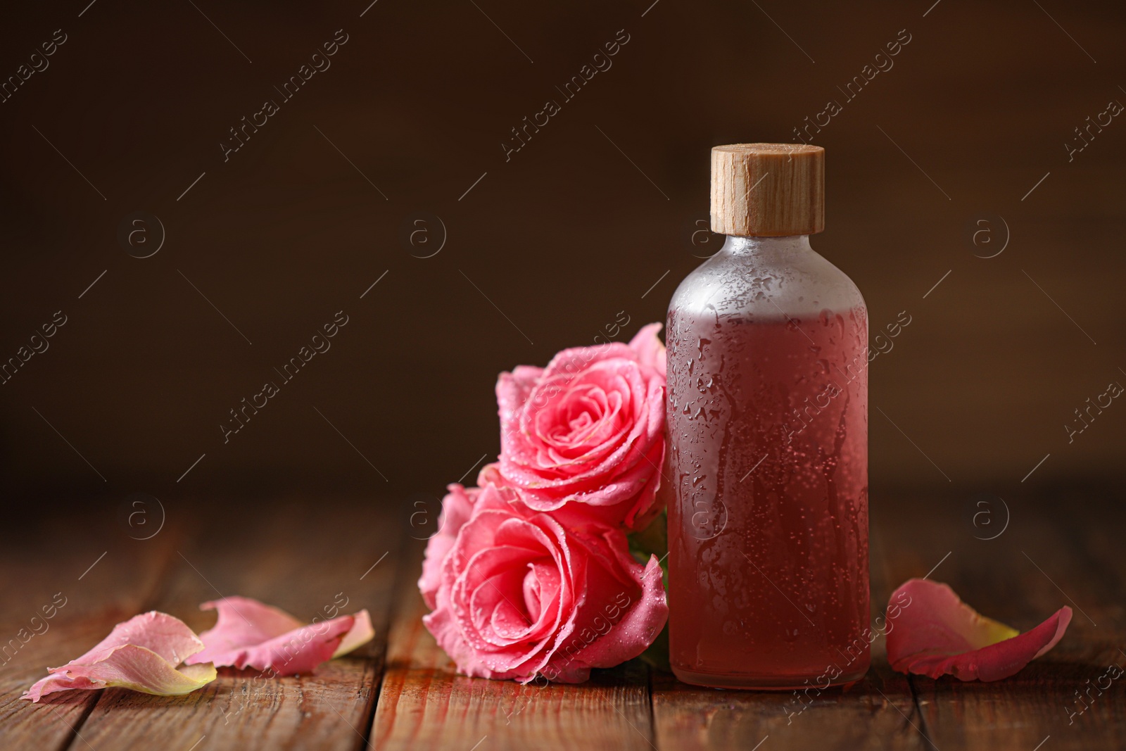 Photo of Bottle of essential rose oil and flowers on wooden table, space for text