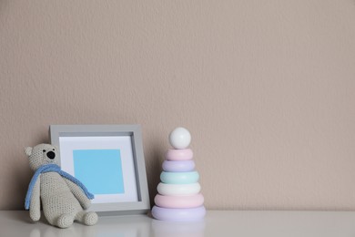 Photo of Empty photo frame, toy bear and ring pyramid on white table near grey wall. Space for design