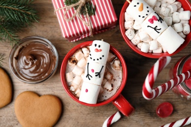 Flat lay composition with funny marshmallow snowmen in cups of hot drinks on wooden table