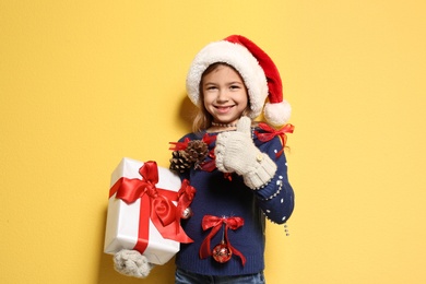 Photo of Cute little girl in handmade Christmas sweater and hat holding gift on color background
