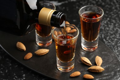 Photo of Pouring tasty amaretto liqueur into glass and almonds on black table, closeup