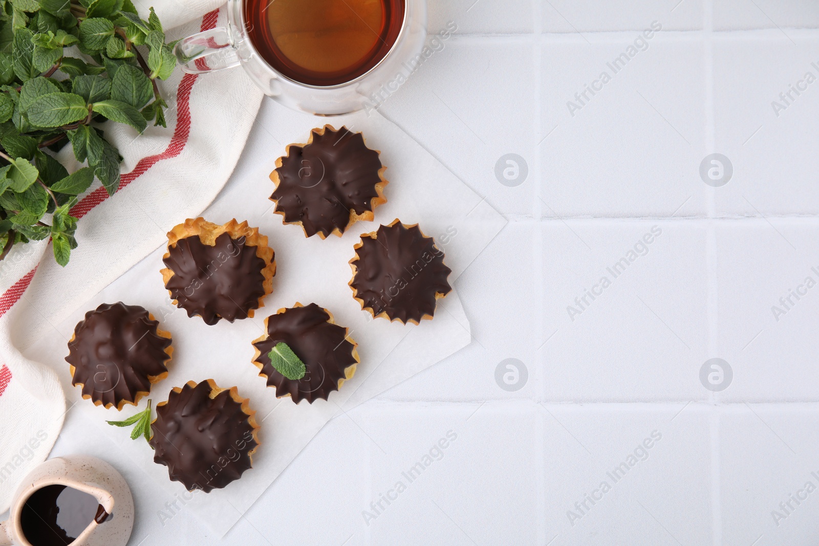 Photo of Delicious profiteroles with chocolate spread, mint and aromatic tea on white tiled table, flat lay. Space for text