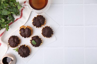 Delicious profiteroles with chocolate spread, mint and aromatic tea on white tiled table, flat lay. Space for text