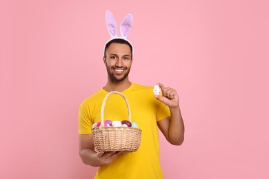 Happy African American man in bunny ears headband holding wicker basket with Easter eggs on pink background