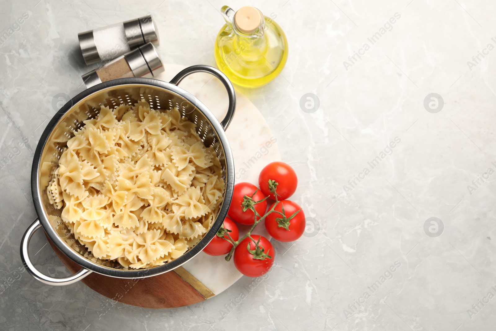 Photo of Cooked pasta in metal colander, spices, oil and tomatoes on grey marble table, flat lay. Space for text