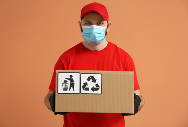 Photo of Courier in mask holding cardboard box with different packaging symbols on orange background. Parcel delivery