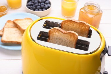 Photo of Yellow toaster with roasted bread, glasses of juice, blueberries and jam on white wooden table, closeup