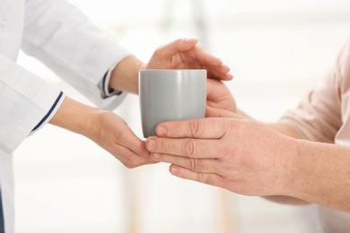 Nurse giving cup of tea to elderly man against blurred background, closeup. Assisting senior generation