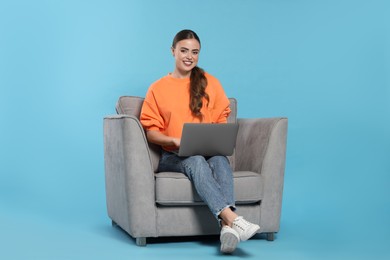 Happy woman with laptop sitting in armchair on light blue background