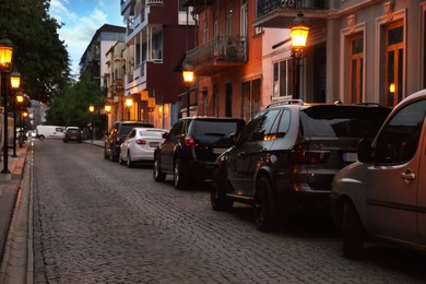 Photo of Glowing streetlights and parked cars on cobblestone road near residential buildings in evening