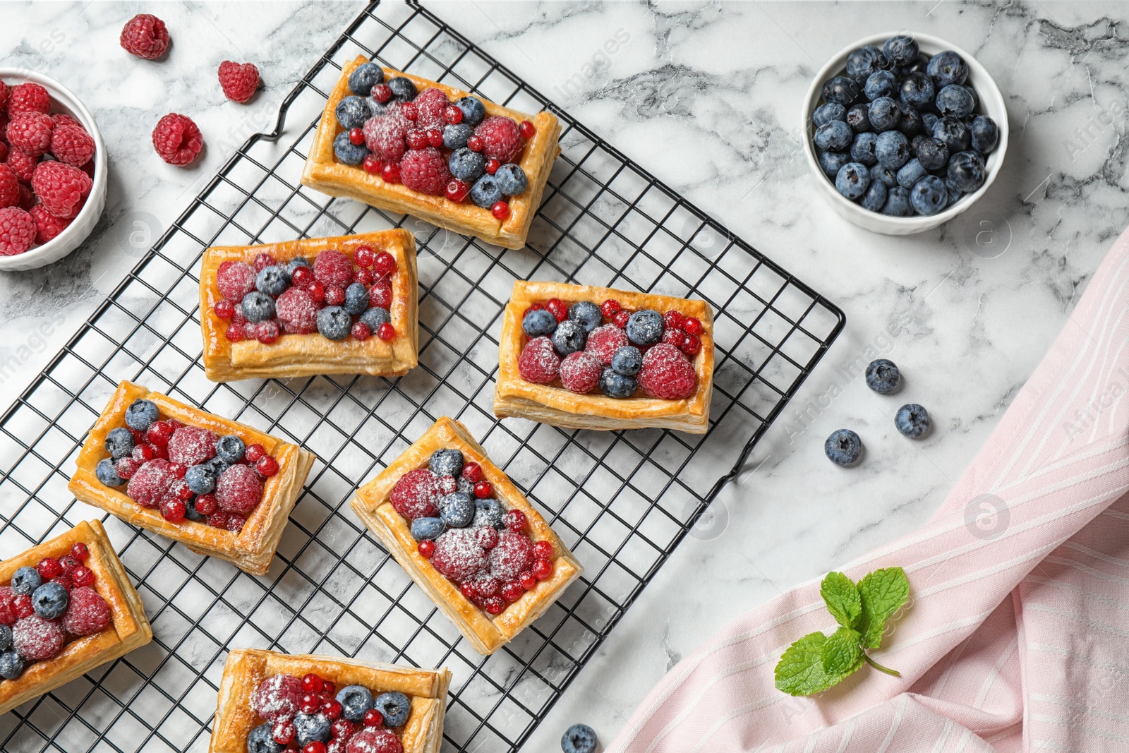 Photo of Cooling rack and fresh delicious puff pastry with sweet berries on white marble table, flat lay