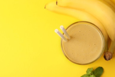 Photo of Glass of tasty banana smoothie with straws, fresh fruits and mint on yellow background, flat lay. Space for text