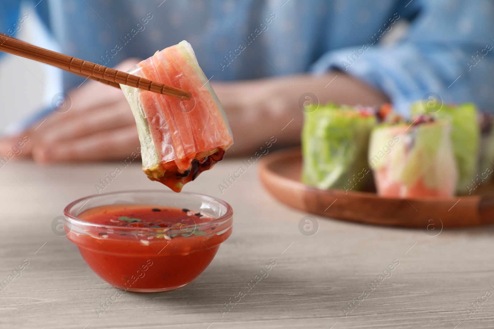 Photo of Woman dipping delicious spring roll wrapped in rice paper into sauce at white wooden table, closeup