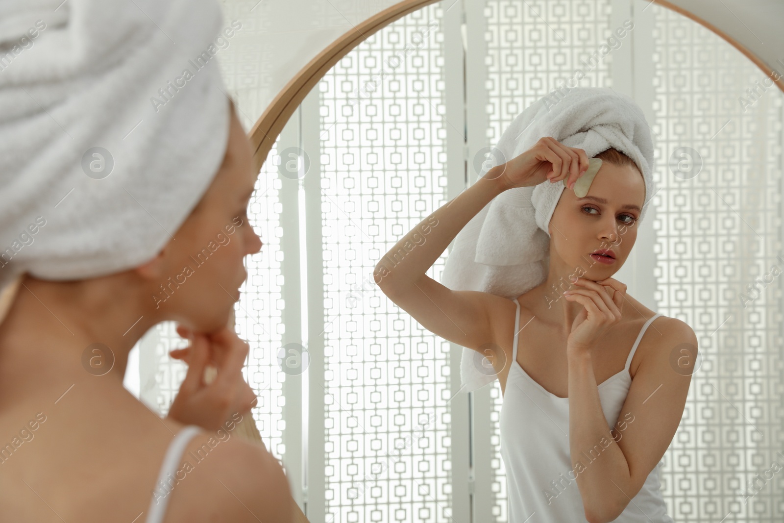 Photo of Beautiful young woman doing facial massage with gua sha tool in front of mirror at home