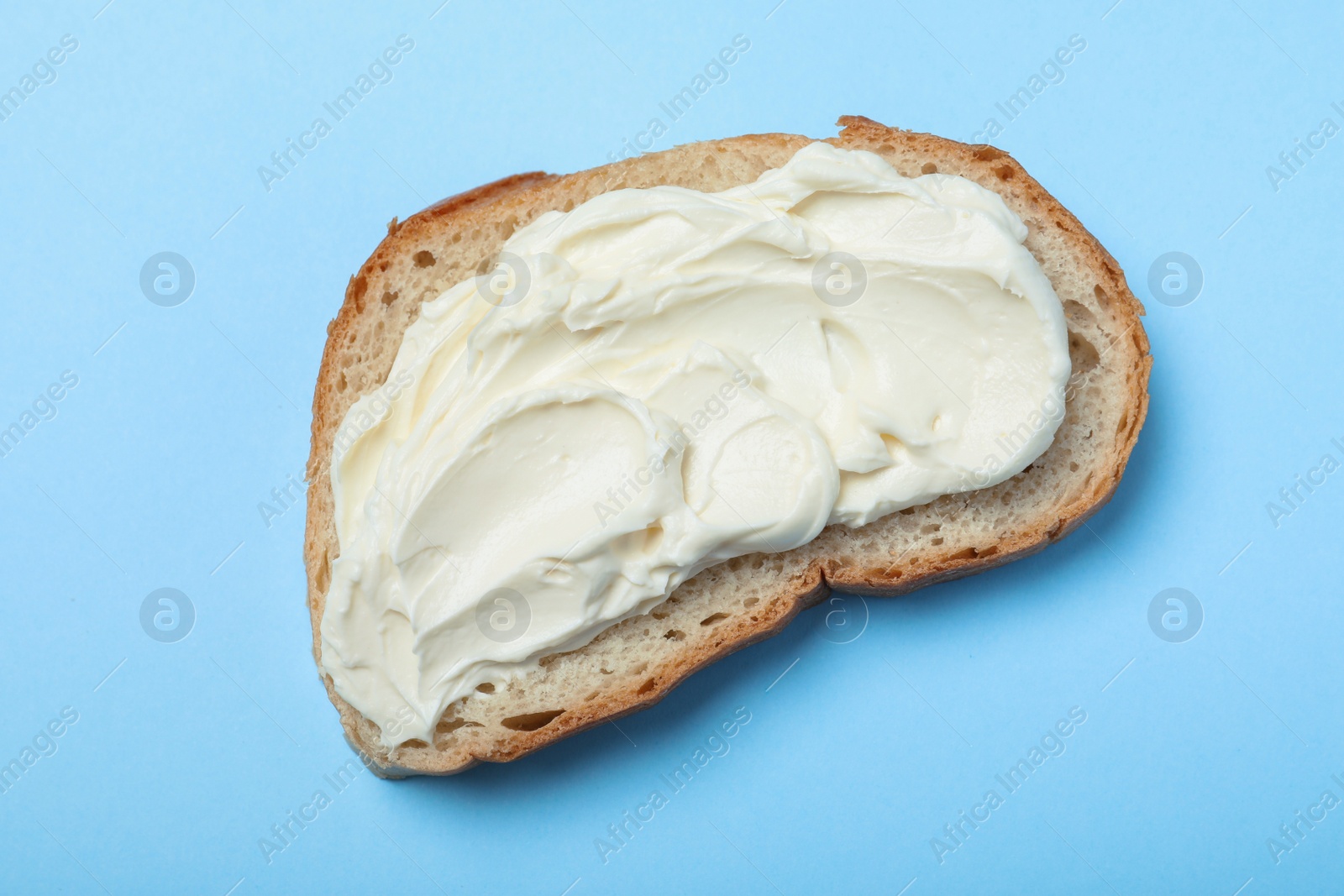 Photo of Slice of bread with tasty cream cheese on light blue background, top view