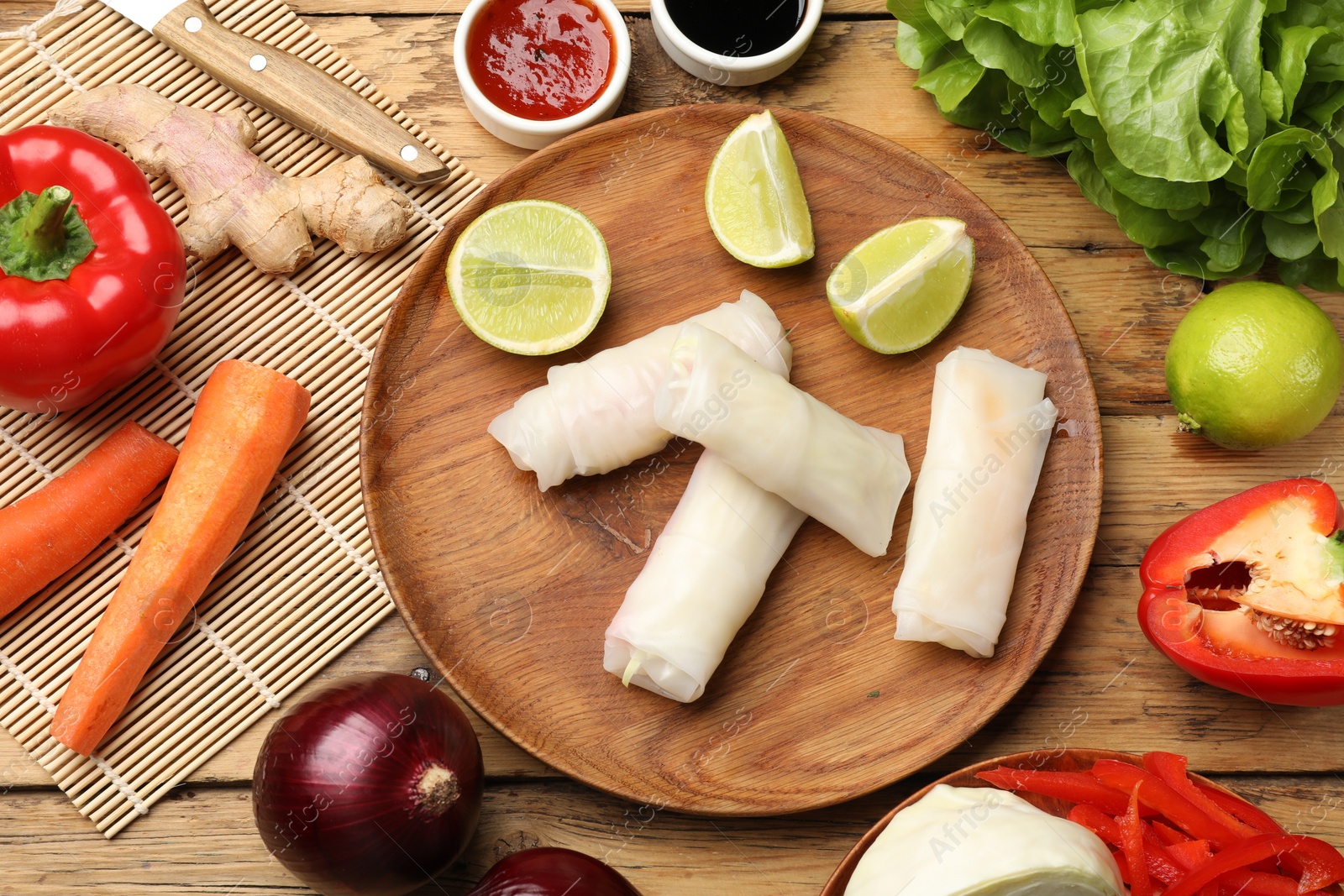 Photo of Delicious spring rolls with vegetables, lime and sauces on wooden table, flat lay