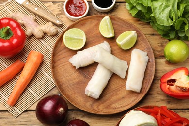 Delicious spring rolls with vegetables, lime and sauces on wooden table, flat lay