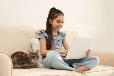 Photo of Cute little girl with laptop and cat on sofa at home. First pet