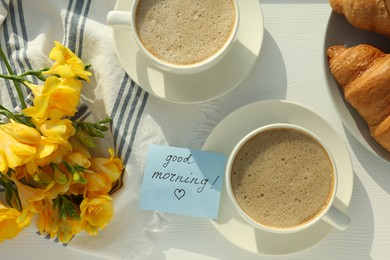 Photo of Coffee, croissants, flowers and card with phrase GOOD MORNING! on white wooden table, flat lay