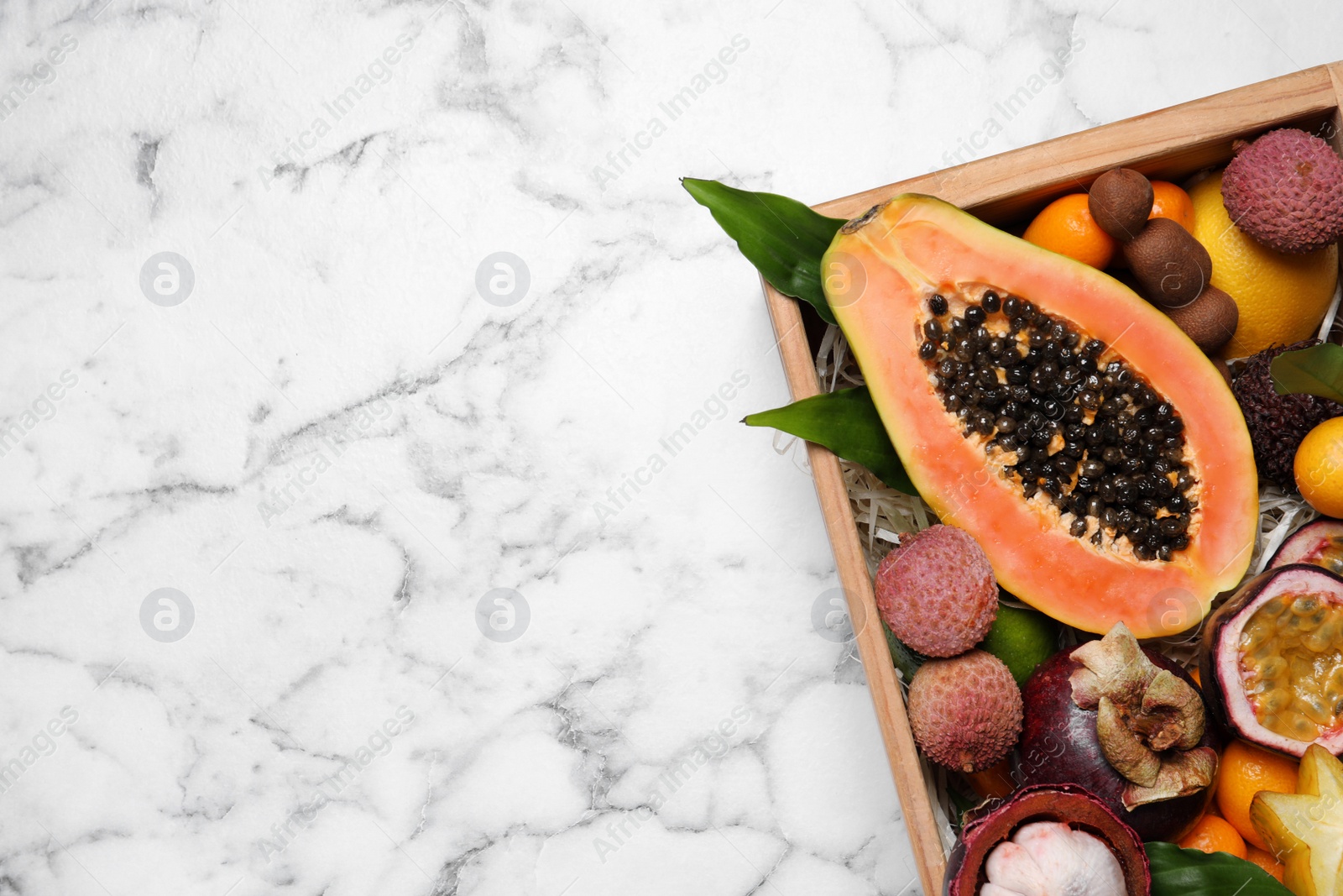 Photo of Different tropical fruits in wooden box on white marble table, top view. Space for text