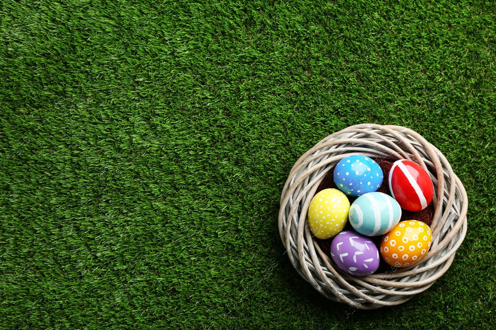 Photo of Wicker nest with painted Easter eggs on green grass, top view. Space for text