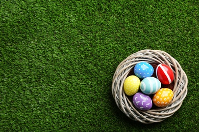 Wicker nest with painted Easter eggs on green grass, top view. Space for text