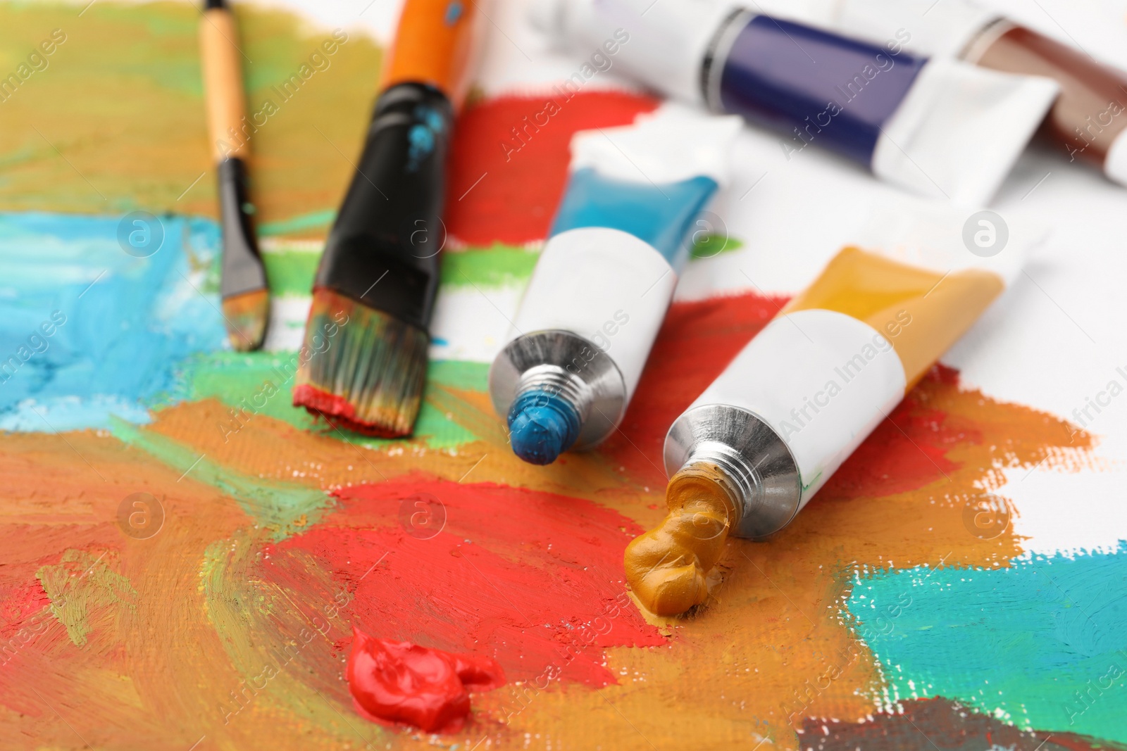 Photo of Tubes of colorful oil paints and brushes on canvas with abstract painting, closeup