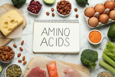 Photo of Different fresh products and notebook with words Amino Acids on white marble table, flat lay